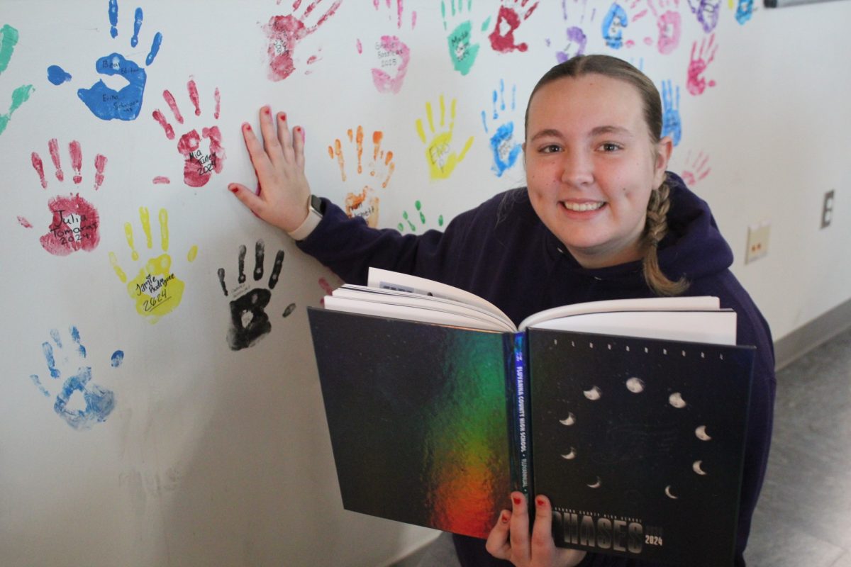 Senior Katie Bond poses next to her handprint on the journalism classroom wall, holding one of the yearbooks she edited.