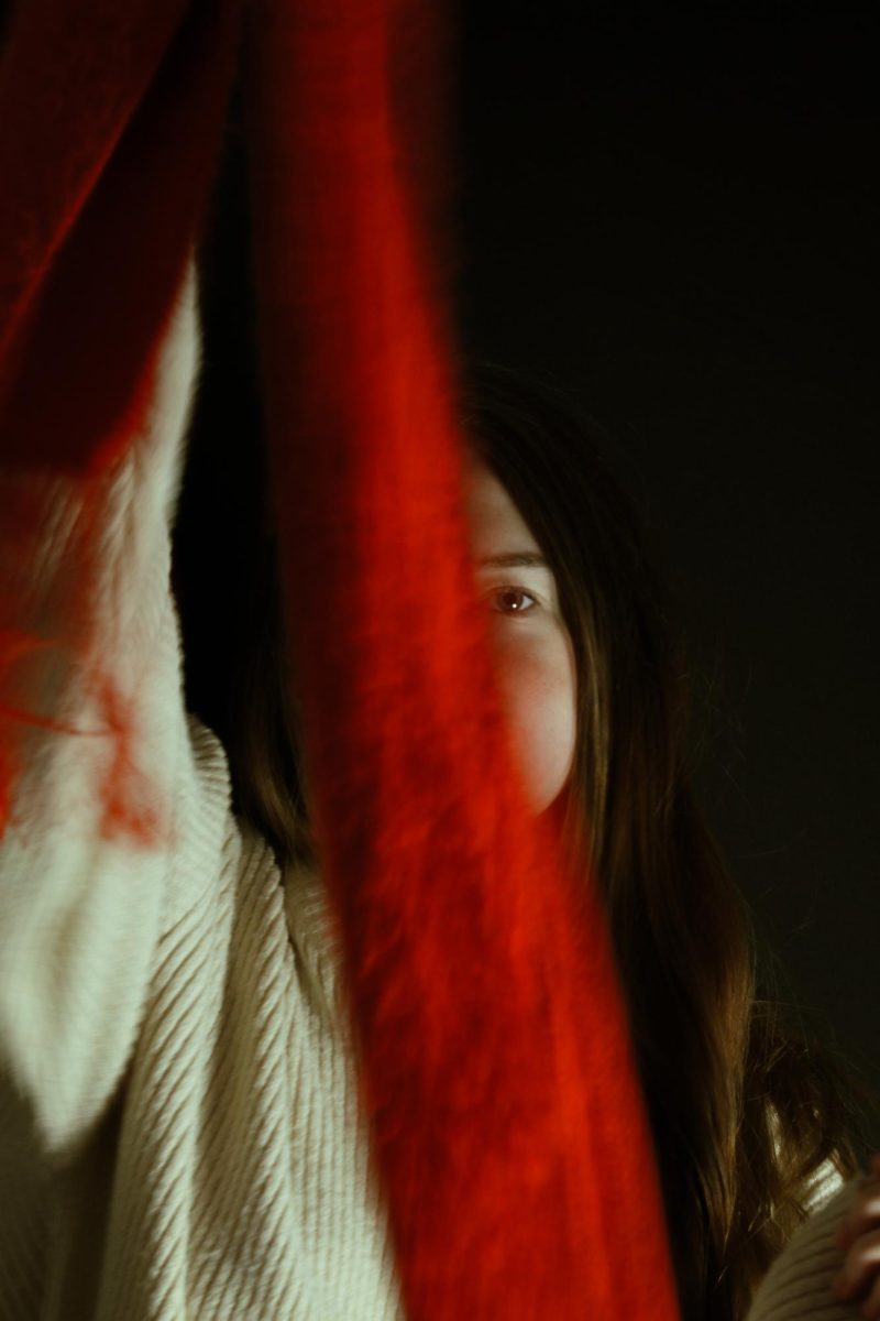 Ella-Grace Mann '27 holding out a red scarf the cover most of her face, leaving only one eye showing, she posed against a black wall. 